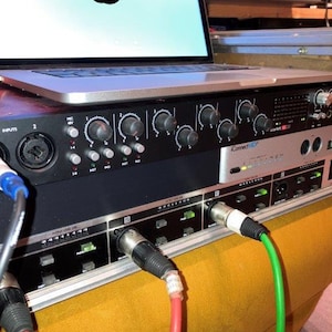 May include: A black audio interface with knobs and buttons, connected to a laptop computer with a silver case. Two cables, one red and one green, are plugged into the interface.