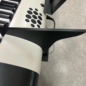 Könnte beinhalten: Eine weiße digitale Tastatur mit einem schwarzen Ständer und einem schwarzen Bildschirm, der oben angebracht ist. Die Tastatur hat ein schwarz-weißes Design und ein Lautsprechergitter an der Seite.