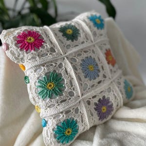 May include: A white crocheted cushion with colourful flower designs and buttons. The cushion is made with a granny square pattern and features a variety of colours, including pink, green, blue, and purple.