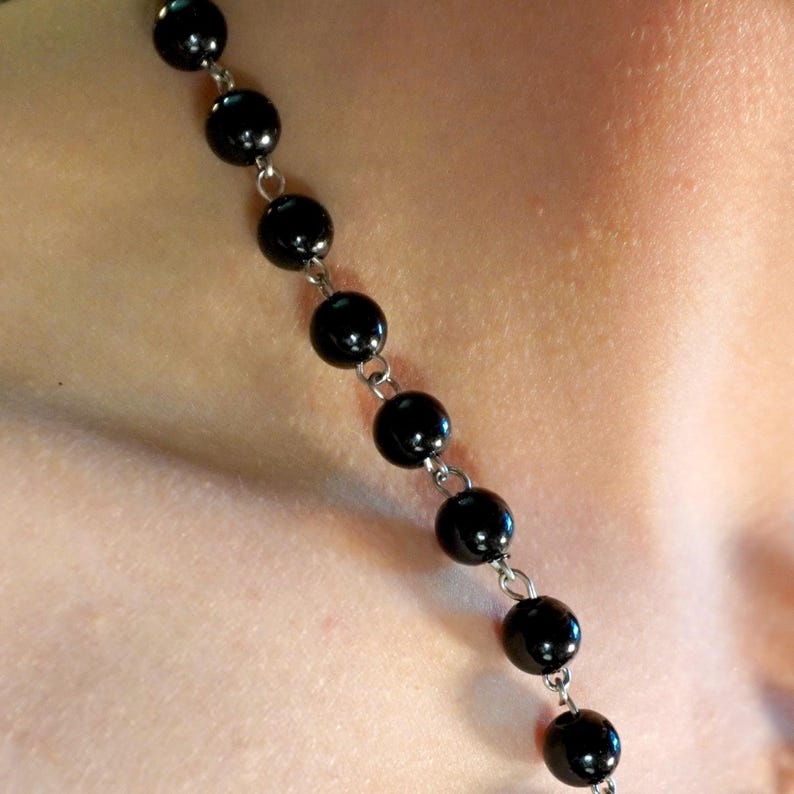 May include: A black beaded necklace with round beads and silver-colored chain links. The necklace is shown against a skin-toned background, suggesting it is being worn. The beads appear to be glossy and evenly spaced.