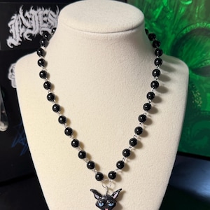 May include: A black beaded necklace with a pendant featuring a black cat with blue eyes and a pink tongue. The cat pendant is attached to a silver-toned metal ring. The necklace is displayed on a white mannequin.