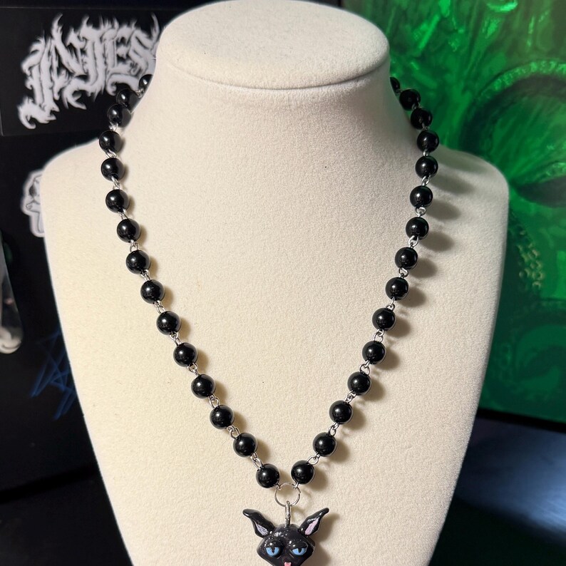 May include: A black beaded necklace with a pendant featuring a black cat with blue eyes and a pink tongue. The cat pendant is attached to a silver-toned metal ring. The necklace is displayed on a white mannequin.