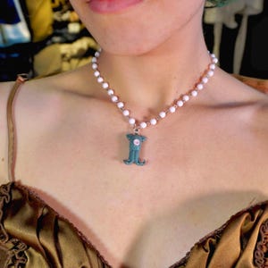 Peut inclure: Un collier compos&eacute; d'une cha&icirc;ne de perles blanches et d'un pendentif turquoise. Le pendentif a un centre rose et une forme stylis&eacute;e unique. Le collier est port&eacute; autour du cou.
