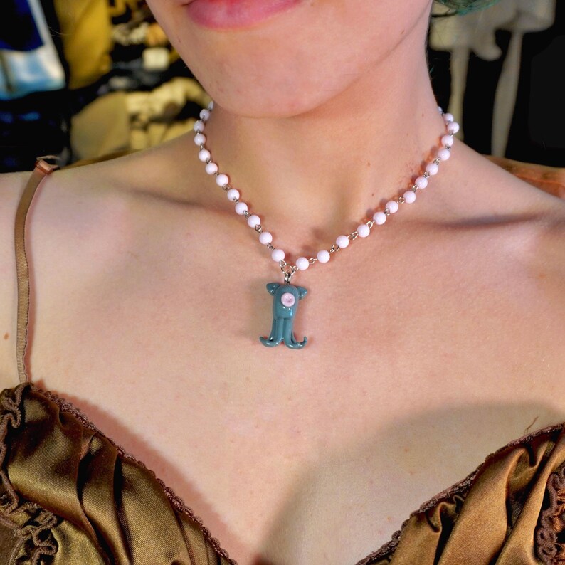 Peut inclure: Un collier compos&eacute; d'une cha&icirc;ne de perles blanches et d'un pendentif turquoise. Le pendentif a un centre rose et une forme stylis&eacute;e unique. Le collier est port&eacute; autour du cou.