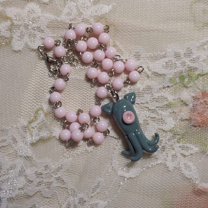 Peut inclure: Un collier avec un pendentif poulpe bleu clair avec un accent rose, et une cha&icirc;ne de perles roses. Le collier est en m&eacute;tal et en verre, et est pr&eacute;sent&eacute; sur une surface en dentelle.