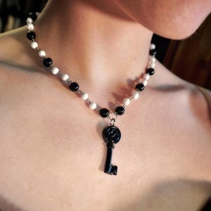 May include: A necklace featuring a black key pendant suspended from a chain of alternating black and white beads. The key has a decorative design on its head. The beads are small and round, creating a contrasting pattern.