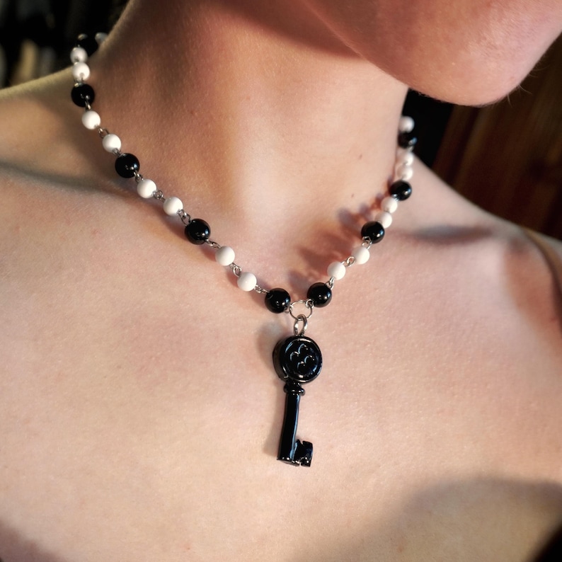 May include: A necklace featuring a black key pendant suspended from a chain of alternating black and white beads. The key has a decorative design on its head. The beads are small and round, creating a contrasting pattern.