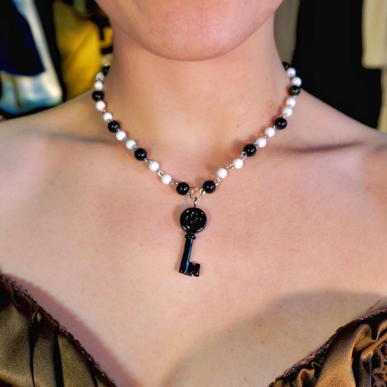 May include: A beaded necklace with alternating white and black beads. A black key pendant hangs from the center. The necklace is worn around the neck.