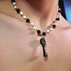 May include: A necklace featuring alternating black and white beads. A black key pendant hangs from the necklace. The key has a decorative design on its head. The necklace is worn around the neck.