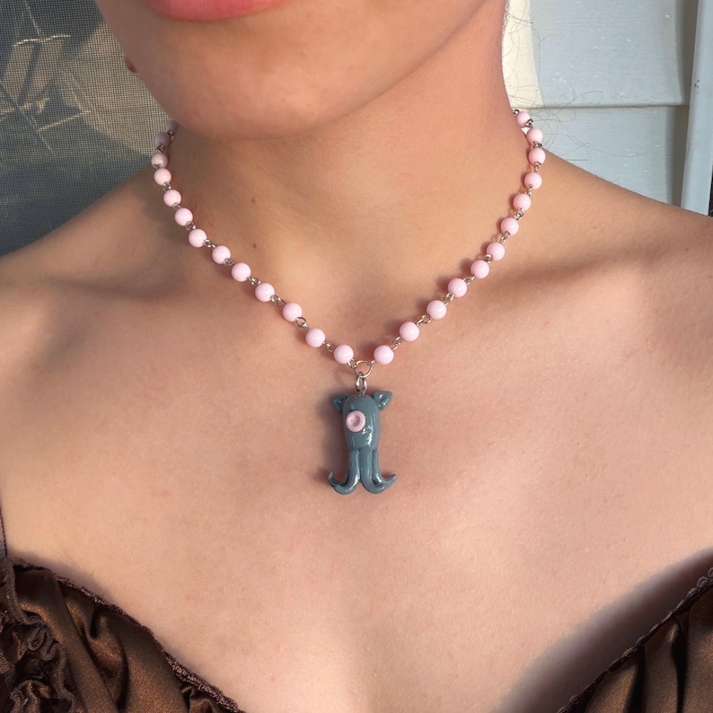 Peut inclure: Un collier de perles roses avec une cha&icirc;ne argent&eacute;e et un pendentif poulpe bleu-vert. Le pendentif poulpe a un &oelig;il rose et une petite boucle pour la cha&icirc;ne. Un bijou d&eacute;licat.