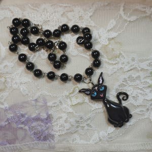 May include: Black beaded necklace with a black cat pendant. The cat pendant has blue eyes and a pink tongue. The necklace is made of small, round, black beads strung on a silver chain.