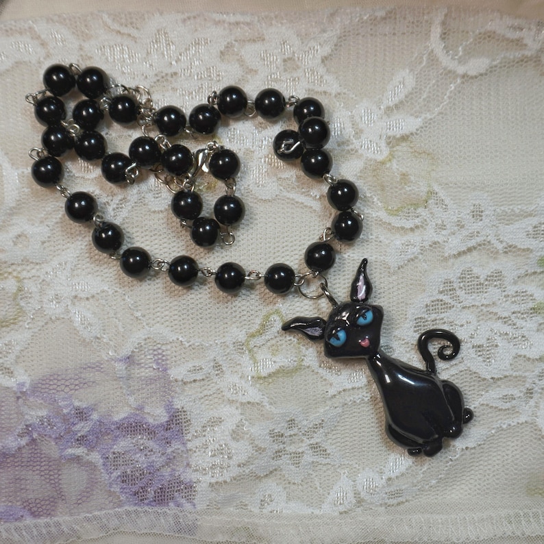 May include: Black beaded necklace with a black cat pendant. The cat pendant has blue eyes and a pink tongue. The necklace is made of small, round, black beads strung on a silver chain.