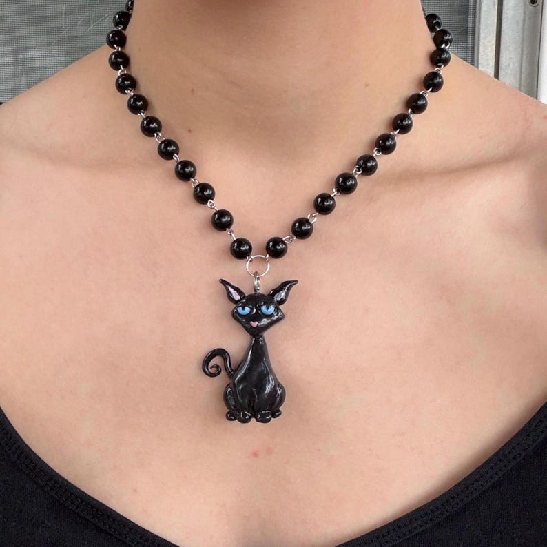 May include: Black beaded necklace with a black cat pendant. The cat pendant has blue eyes and a curled tail. The necklace is worn around the neck.