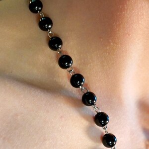 May include: A black beaded necklace with round beads and silver-colored chain links. The necklace is shown against a skin-toned background, suggesting it is being worn. The beads appear to be glossy and evenly spaced.