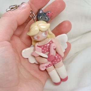 May include: A handmade keychain featuring a Cupid figurine with blonde hair, white wings, and a pink polka-dot outfit. A gray cat with a pink bow sits atop the Cupid's head. The keychain is attached to a silver chain.