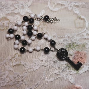 May include: A necklace featuring alternating black and white beads, connected by a silver chain. A black key pendant hangs from the necklace. The necklace is displayed on a white lace surface.
