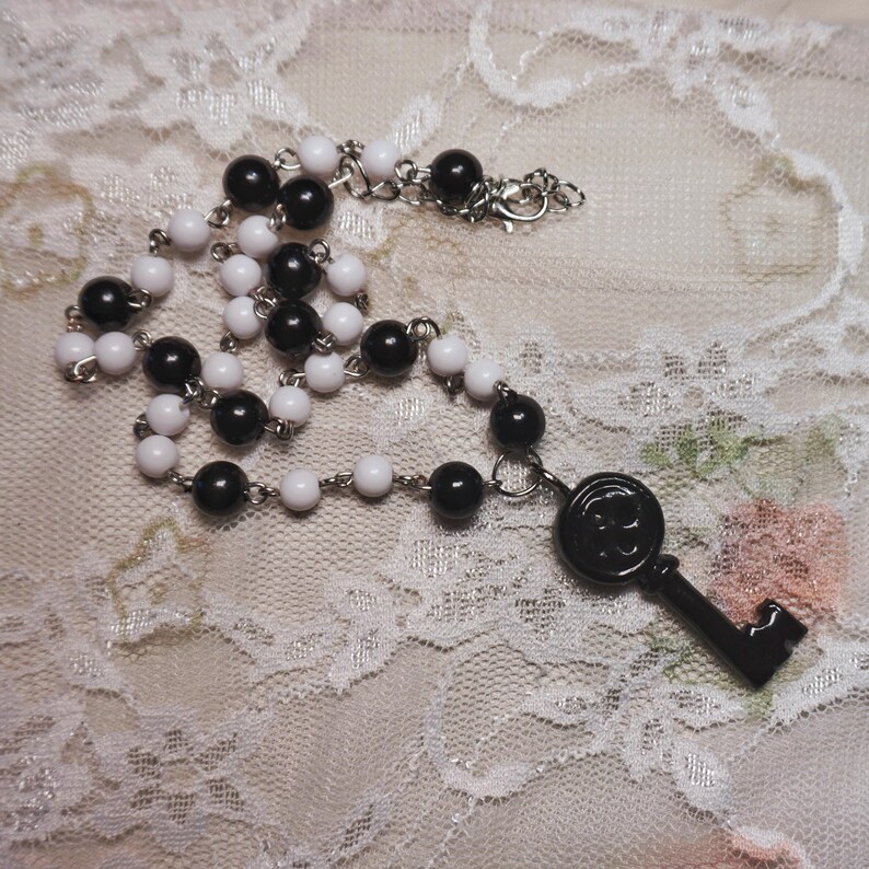 May include: A necklace featuring alternating black and white beads, connected by a silver chain. A black key pendant hangs from the necklace. The necklace is displayed on a white lace surface.