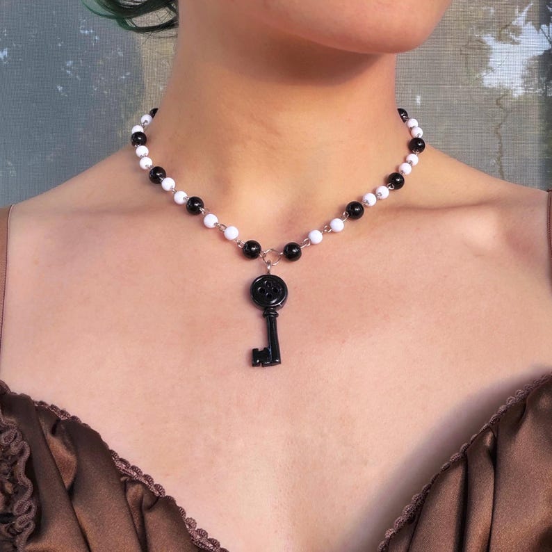 May include: A necklace featuring a black key pendant suspended from a strand of alternating white and black beads. The key has a decorative top and a detailed handle. The necklace is worn around the neck.