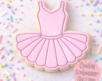 Op de afbeelding: Een roze koekje in de vorm van een ballerina tutu jurk. De koek heeft een lichtroze lijfje en een gerimpelde rok. De koek is versierd met een lichtroze omtrek en ligt op een wit oppervlak met kleurrijke hagelslag. Tekst op de afbeelding zegt "Cookie Embosser By Lisa."