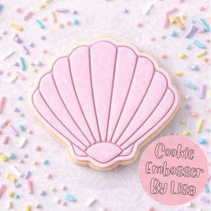 May include: A pink seashell-shaped biscuit with a light-coloured outline and a textured surface. The biscuit is decorated with pink icing and surrounded by colourful sprinkles. The text "Cookie Embosser By Lisa" is in a pink circle.