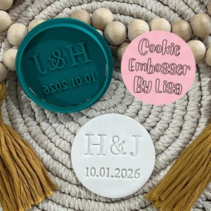 May include: A selection of cookie embossers in different colours and designs. One teal embosser has the letters "L & H" and the date "10.01.2026". A pink embosser reads "Cookie Embosser By Lisa". A white embosser displays "H & J" and the date "10.01.2026".