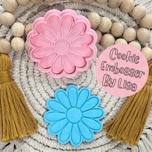 May include: Two daisy-shaped cookie cutters, one pink and one blue, are displayed. The pink cutter is on top, and the blue one is below. A pink circle with the text "Cookie Embosser By Lisa" is also visible. The background includes wooden beads and tassels.