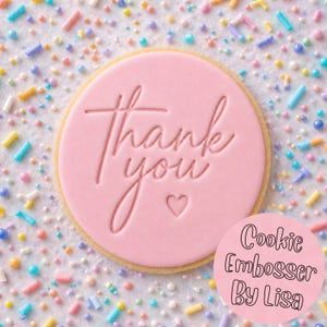 May include: A round cookie with a pink embossed design that says "Thank You" with a heart. The cookie is surrounded by colourful sprinkles. The text "Cookie Embosser By Lisa" is in a pink circle.