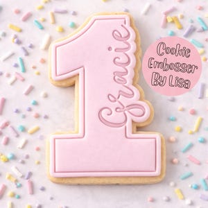 May include: A pink number one cookie with the name "Gracie" in cursive script. The cookie has a pink frosting outline and a scalloped edge. The text "Cookie Embosser By Lisa" is in a pink circle. The cookie is surrounded by colourful sprinkles.