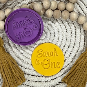 May include: Two round cookie cutters, one purple and one yellow. The purple cutter is for making a cookie with the text "Sarah is One" in a cursive font. The yellow cutter is for making a cookie with the text "Sarah is One" in a cursive font.