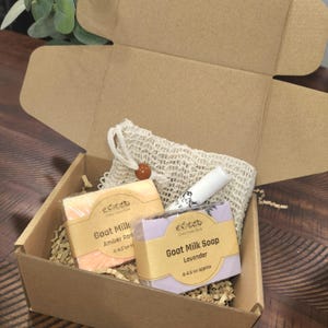 Puede incluir: Una caja de cartón marrón con una bolsa de malla blanca, dos barras de jabón de leche de cabra y un bálsamo labial. Las barras de jabón están etiquetadas como "Goat Milk Amber Rose" y "Goat Milk Soap Lavender".