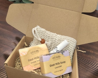 May include: A brown cardboard box with a white mesh bag, two bars of goat milk soap, and a lip balm. The soap bars are labeled "Goat Milk Amber Rose" and "Goat Milk Soap Lavender".