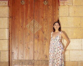 May include: A woman wearing a white maxi dress with a floral pattern stands in front of a wooden door with ornate brass accents. The door is set in a stone archway.