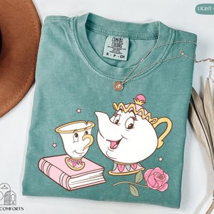 May include: Light green Comfort Colors t-shirt featuring a cartoon illustration of a teapot and teacup with a pink rose. The teapot and teacup are smiling. A gold necklace is draped across the shirt. The shirt is labeled "S-P-CH".