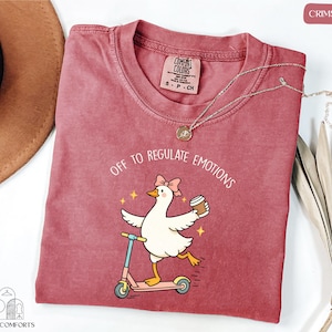 A red t-shirt with a cartoon image of a duck riding a scooter, accompanied by the text "Off to regulate emotions".
