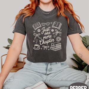 A woman wearing a gray t-shirt with a graphic design and text printed on it, sitting on a chair with plants in the background.