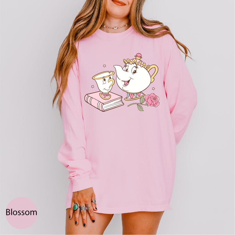 A woman wearing a pink long-sleeved shirt with a cartoon illustration of a teapot and a book on it.