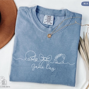 May include: A blue jean Comfort Colors t-shirt with a white graphic of a football helmet, bow, and football, with the words "Game Day" underneath. A gold necklace with a pendant is on the shirt.