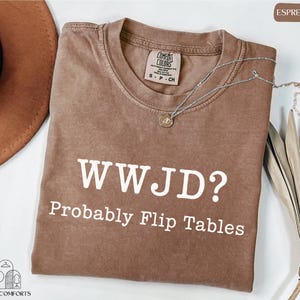 a brown t-shirt with the text "WWJD?" printed on it, along with the phrase "Probably Flip Tables" below it. The t-shirt is laid out on a white surface, and there is a hat and some leaves nearby.