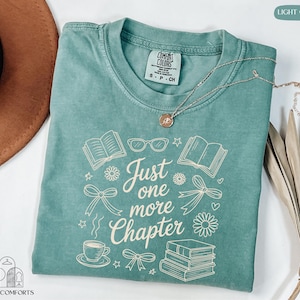 A turquoise t-shirt with the text "Just one more Chapter" printed on it, along with various book-related icons and illustrations. The t-shirt is folded and placed on a white surface, with a brown hat and a plant nearby.