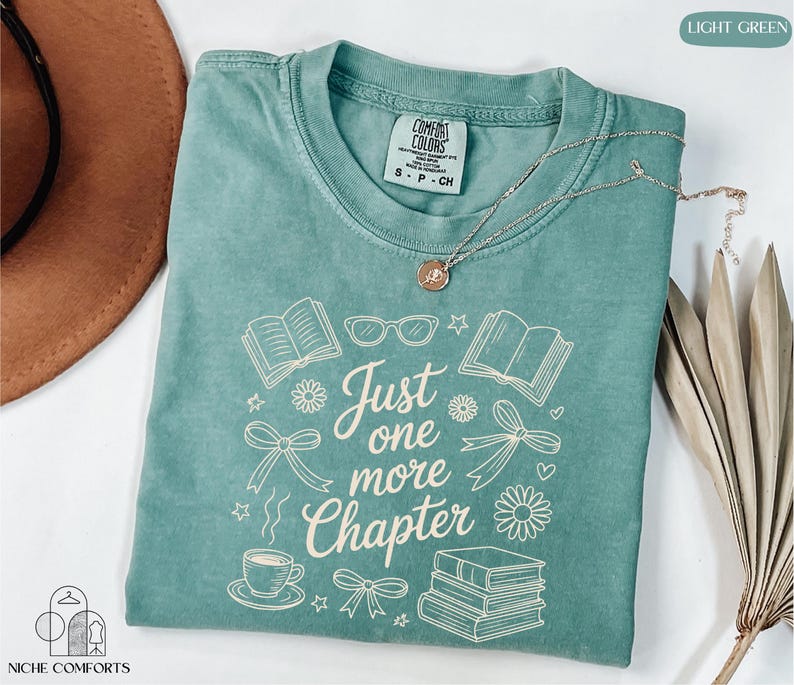 A turquoise t-shirt with the text "Just one more Chapter" printed on it, along with various book-related icons and illustrations. The t-shirt is folded and placed on a white surface, with a brown hat and a plant nearby.