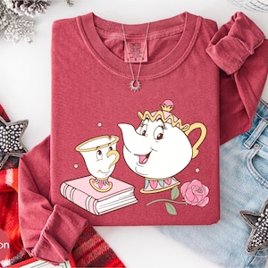 A red t-shirt with a cartoon illustration of a teapot and a character from the Disney film Beauty and the Beast, along with a necklace and a pair of jeans, are displayed on a white background.