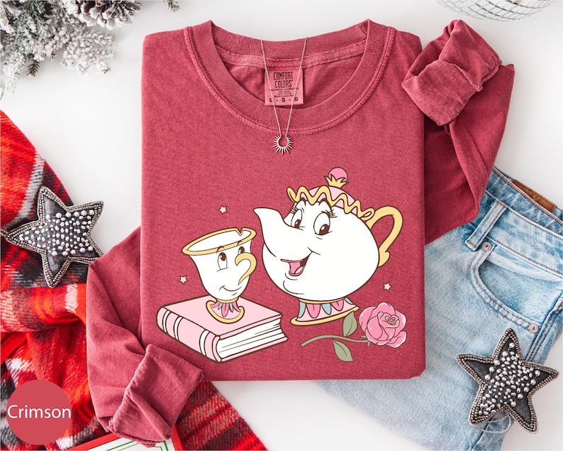 A red t-shirt with a cartoon illustration of a teapot and a character from the Disney film Beauty and the Beast, along with a necklace and a pair of jeans, are displayed on a white background.