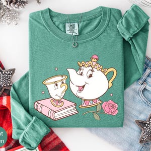 a green long-sleeved shirt with a cartoon illustration of a teapot and a cup, along with a rose and a book, all set against a white background.