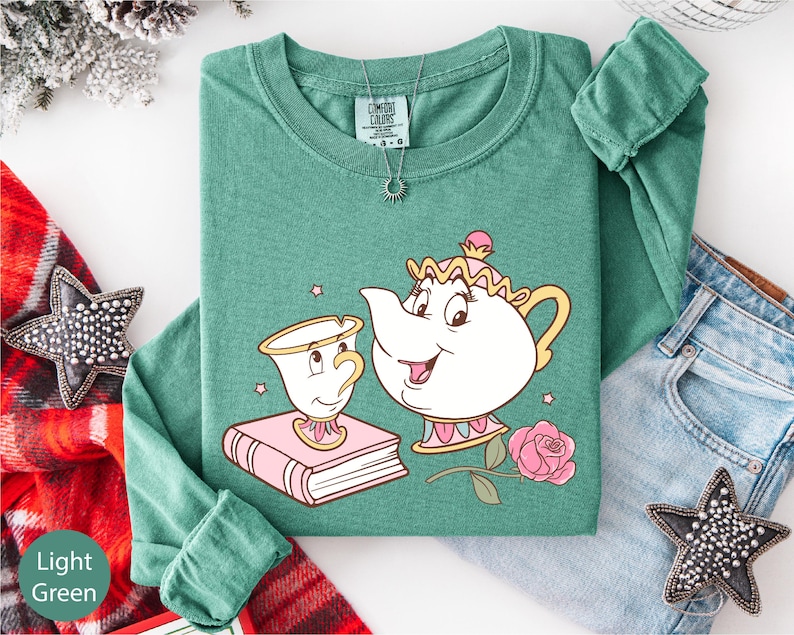 a green long-sleeved shirt with a cartoon illustration of a teapot and a cup, along with a rose and a book, all set against a white background.