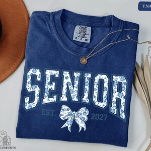 a blue t-shirt with the text "SENIOR" and a bow, along with the text "EST. 2027" below it. The t-shirt is laid out on a white surface, and there is a brown hat and some dried leaves nearby.