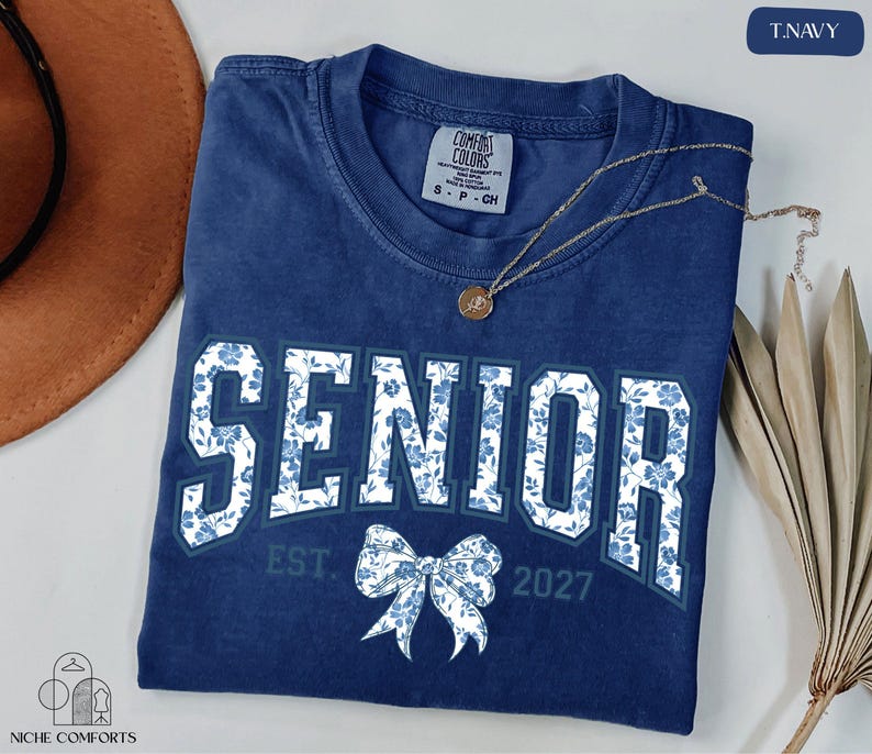 a blue t-shirt with the text "SENIOR" and a bow, along with the text "EST. 2027" below it. The t-shirt is laid out on a white surface, and there is a brown hat and some dried leaves nearby.