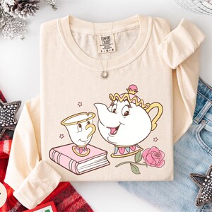 A beige long-sleeved t-shirt with a cartoon illustration of a teapot and a cup, along with a necklace and a book, is displayed on a white background.