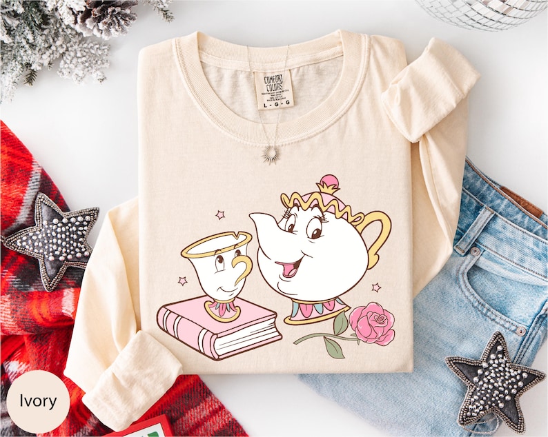 A beige long-sleeved t-shirt with a cartoon illustration of a teapot and a cup, along with a necklace and a book, is displayed on a white background.