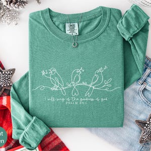 a green long-sleeved shirt with a hand-drawn design of three birds and the text "I will sing of the glory of God" below the design. The shirt is laid out on a white surface, accompanied by a pair of jeans, a red and white plaid blanket, and some decorative star-shaped embellishments.