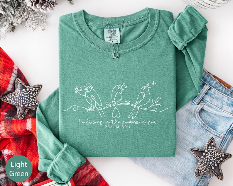 a green long-sleeved shirt with a hand-drawn design of three birds and the text "I will sing of the glory of God" below the design. The shirt is laid out on a white surface, accompanied by a pair of jeans, a red and white plaid blanket, and some decorative star-shaped embellishments.